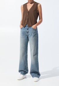 Personne portant un gilet marron boutonné, un jean large bleu clair, des baskets blanches, debout les mains dans les poches sur fond blanc.