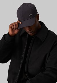 Casquette de baseball noire avec le logo "CK", en tissu texturé. Portée avec une veste noire zippée à col haut.