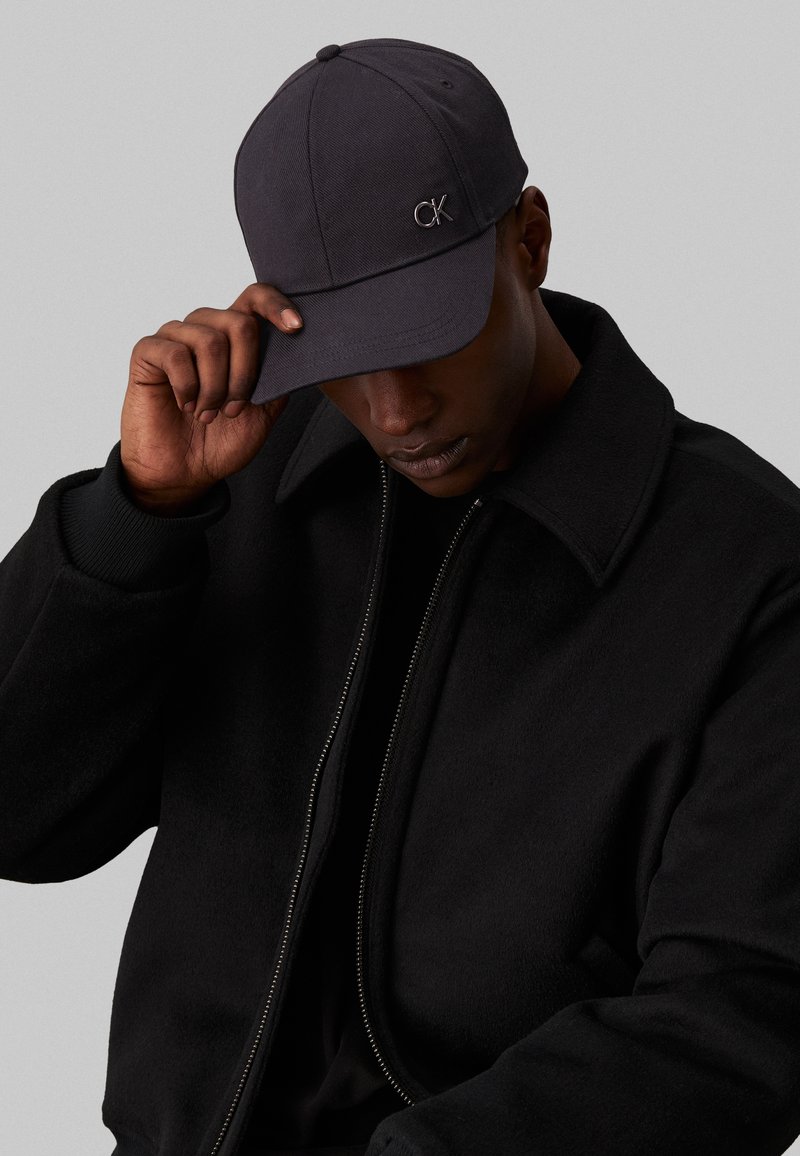 Casquette de baseball noire avec le logo "CK", en tissu texturé. Portée avec une veste noire zippée à col haut.