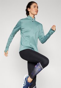 Licht teal lange mouw sporttop met een ritskraag, gecombineerd met zwarte legging met een kleurrijke patroonaccent op de onderkant van het been.