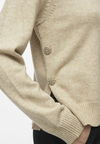 Beez värvi kudum, millel on ribbed varrukad ja küljed, kaunistatud kahe lillekujulisest metallnupuga, pehme tekstuur ja mugav lõige.