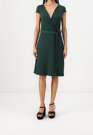 Femme portant une robe portefeuille vert foncé à manches courtes avec ceinture à la taille et des sandales compensées noires à talons hauts sur un fond uni.