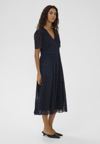 Tengerészkék maxi ruha rövid áttetsző ujjal és V-nyakkivágással. Virágmintás és légies szoknyával, fekete belebújós cipővel párosítva.