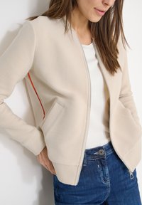 Femme portant une veste beige côtelée à fermeture éclair avec un détail rouge sur le côté, chemise blanche et jean bleu, les mains dans les poches de la veste.