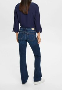 Blouse marine avec manches nouées portée sur un jean évasé bleu foncé. Le jean comporte des poches arrière et une étiquette de marque claire sur la ceinture.