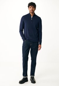 Marineblauwe pullover met een ritskraag, gecombineerd met donkere denim jeans. Het model draagt zwarte enkellaarsjes met een opgerolde zoom bij de broekspijpen.