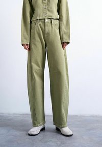 Personne portant un pantalon large taille haute vert olive, une veste boutonnée assortie, des chaussures blanches à talons et des bagues aux doigts, debout sur un sol en béton.