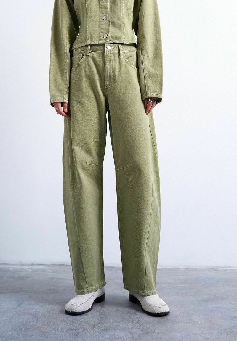 Personne portant un pantalon large taille haute vert olive, une veste boutonnée assortie, des chaussures blanches à talons et des bagues aux doigts, debout sur un sol en béton.