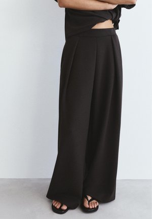 Osoba oblečená v čierne široké nohavice a čierne sandále, stojaca s rukami založenými na jednoduchom svetlom pozadí.