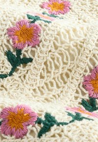 Couverture en coton brodée avec une base crème ornée de motifs floraux en rose et jaune, rehaussée de tiges vertes. Design texturé semblable à de la dentelle.