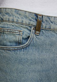 Helle blaue Jeans mit Metallknopfverschluss, sichtbaren Nähakzenten und einer kleinen Fronttasche nahe dem Bund.