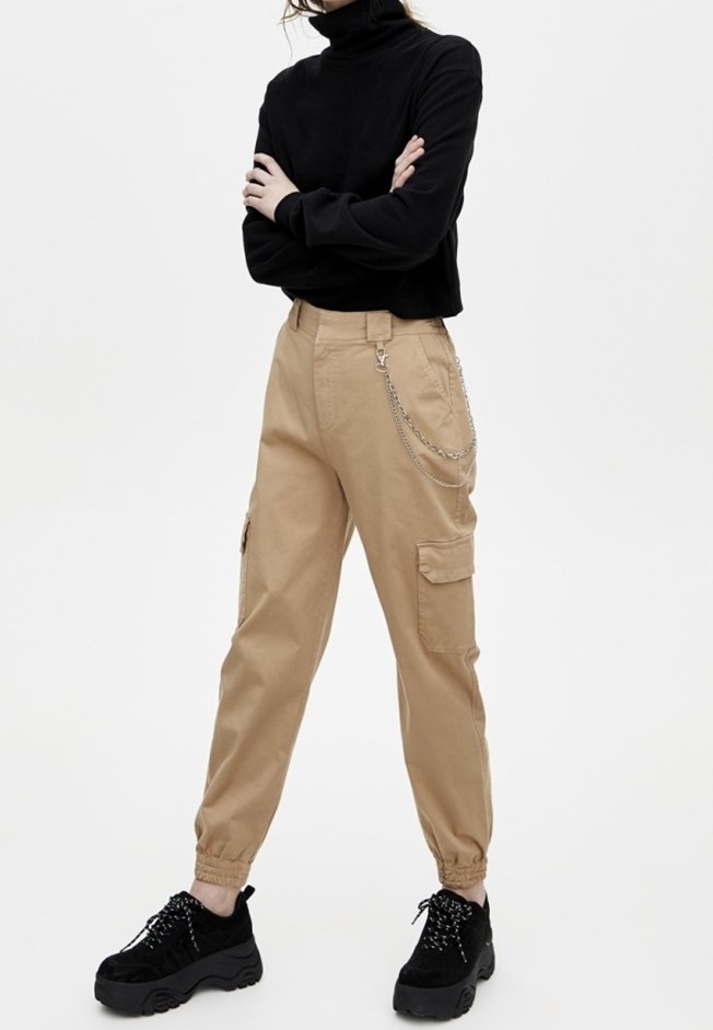 Personne portant un pantalon cargo beige avec détail chaîne, un pull col roulé noir et des baskets à plateforme noires, debout les bras croisés.