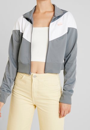 Kobieta nosząca szarą i białą kurtkę Nike na zamek błyskawiczny, na białym krótkim topie i jasnożółtych spodniach z wysokim stanem.