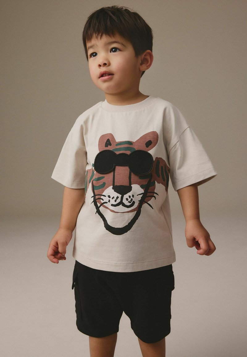 Junges Kind trägt ein beiges T-Shirt mit großem Tigerkopf und 3D Sonnenbrillendesign, kombiniert mit schwarzen Shorts, steht vor einfachem Hintergrund.