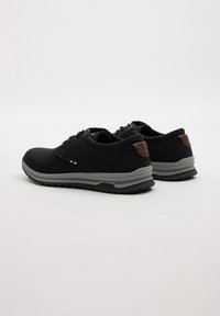 Zapatillas deportivas negras con suela gris, parte superior texturizada, cordones planos y un acento marrón en el talón. Cuentan con un diseño liso y acolchado.