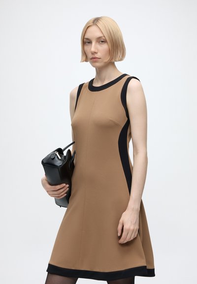 Femme aux cheveux courts et blonds portant une robe beige sans manches avec un galon noir, tenant un sac à main noir sur un fond clair uni.