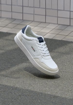 White sneaker with a smooth leather upper, textured suede accents, navy heel tab, and lace closure, displayed against a grey pavement background.
