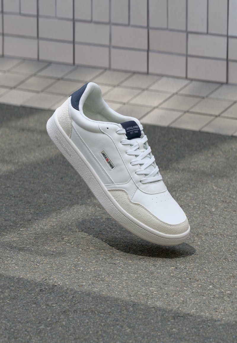 White sneaker with a smooth leather upper, textured suede accents, navy heel tab, and lace closure, displayed against a grey pavement background.