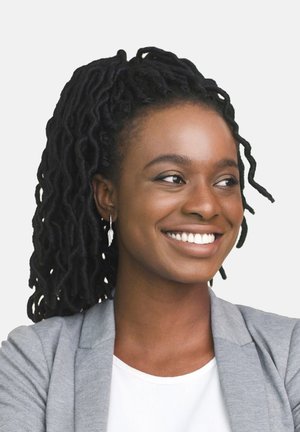 Una donna con lunghi dreadlocks neri indossa un blazer grigio chiaro sopra una maglietta bianca, presenta un orecchino a forma di piuma argentato e un ampio sorriso.