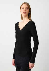 Schwarzes Langarmshirt mit geripptem Muster und V-Ausschnitt, der mit Spitzenbesatz akzentuiert ist. Die Passform ist tailliert und figurbetont.