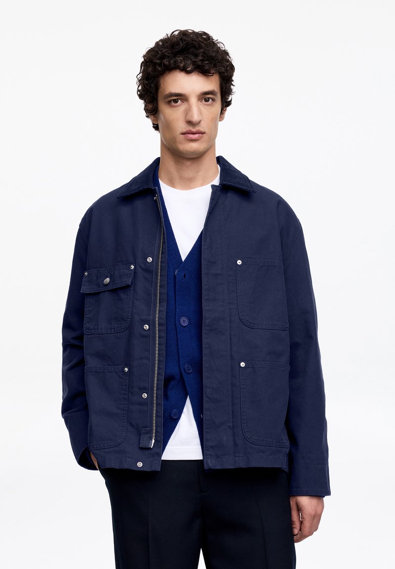 Homme aux cheveux bouclés portant une veste bleu foncé par-dessus un cardigan bleu et un t-shirt blanc, debout devant un fond blanc uni.