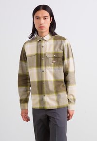 Chemise en flanelle à carreaux dans des nuances de vert olive, beige et jaune. Avec une fermeture à boutons sur le devant et une poche de poitrine avec rabat et bouton-pression.