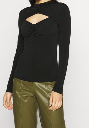 Kobieta ubrana w czarną bluzkę z długim rękawem z marszczonym wycięciem na klatce piersiowej i oliwkowozielone spodnie ze sztucznej skóry, stojąca na jednolitym tle.