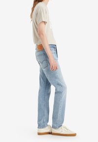 Hellblaue Jeans mit geradem Bein, einer Lederapplikation am Rücken und dezenten Verwaschungen, kombiniert mit einem beigen T-Shirt und weißen Sneakers.