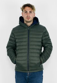Giacca puffa di colore verde scuro con design trapuntato orizzontale, chiusura frontale con zip e cappuccio foderato in blu navy. Due tasche laterali.