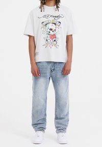Camiseta blanca de manga corta con un gráfico colorido de una calavera y el texto "LOVE DIE HARD". Combinada con vaqueros holgados de color azul claro y zapatillas blancas.