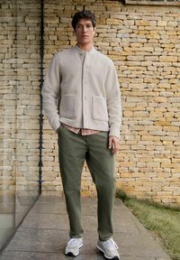 Light grey button-up jacket with two front pockets, worn over a plaid shirt. Olive green pants and grey sneakers complete the outfit.