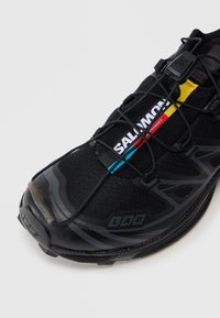 Sapatilha atlética preta com parte superior em malha, apresentando o proeminente logotipo branco "Salomon", detalhes coloridos e uma sola texturizada para aderência.