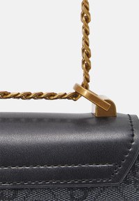 Detailní záběr na černou texturovanou kabelku s černými koženými detaily a zkrouceným zlatým řetízkem připevněným zlatým kováním.