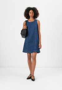 Denim jurk met een mouwloos ontwerp, A-lijn vorm, donkerblauwe kleur, met zichtbare stiksels en een zoom die iets uitloopt. Houdt een zwarte tas vast.
