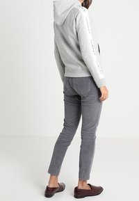 Vrouw die een grijze capuchontrui draagt met de tekst "GANT" op de mouwen, grijze slim-fit broek en donkerbruine loafers, staand met de rug naar de camera.