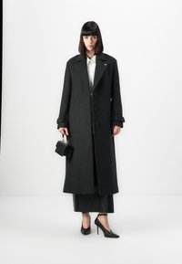 Long black wool coat with a tailored fit, pronounced lapels, and large buttons; paired with a white shirt and sleek black accessories.