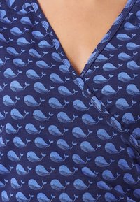 Námořnická modrá tkanina s opakujícím se vzorem světle modrých velryb. Design obsahuje výstřih do V a má jemnou texturu s decentními liniovými akcenty.