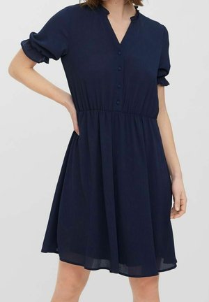 Robe bleu marine à manches courtes avec boutons sur le devant, taille cintrée et manches à volants, portée par une personne debout avec une main sur la hanche.