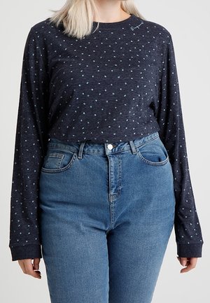 Vrouw met een donkerblauw, met stippen bedrukt, shirt met lange mouwen, ingestopt in een hooggestemde blauwe spijkerbroek, staand tegen een effen achtergrond.