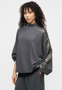 Ασύμμετρο, σκούρο γκρι πουλόβερ από μαλακό ύφασμα. Διαθέτει ψηλό γιακά, άνετη εφαρμογή και διακριτική λεπτομέρεια με λογότυπο μπροστά. Ριγέ τελείωμα.