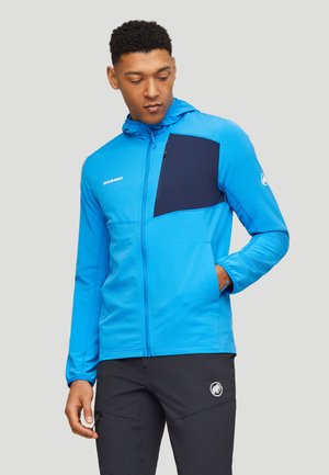 Mann trägt eine blau-schwarze Jacke mit Reißverschluss und Kapuze sowie schwarze Hose und steht mit einer Hand in der Tasche.