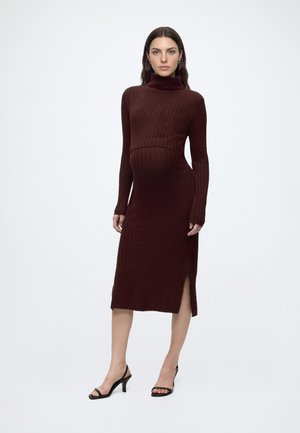 Femme enceinte portant une robe en maille côtelée bordeaux à manches longues avec col roulé et fente sur le côté, associée à des sandales à talons noires.