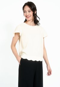 Kreem värvi lühikeste varrukate ja ümarate kaelustega topp, millel on sakiline äärejoon, koos mustade avarate pükstega, mis demonstreerivad sileda ja kerge materjali.