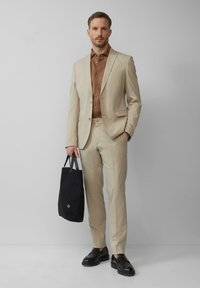 Beige op maat gemaakt pak met een gestructureerde afwerking, gecombineerd met een bruine overhemd. Zwarte instap schoenen en een donkere handtas maken de look compleet.