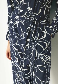 Robe en tissu texturé bleu marine avec des motifs floraux blancs, dotée d'une taille nouée et de manches longues, mettant en valeur une silhouette élégante.