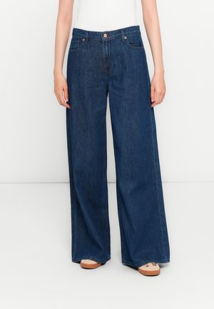 Persoon in donkerblauwe wijde jeans en beige sneakers, staande tegen een eenvoudige witte achtergrond.