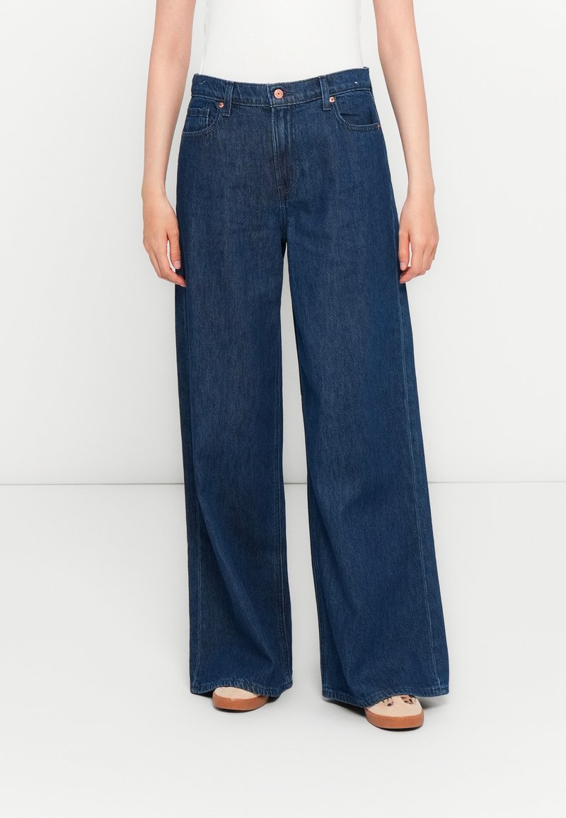 Persoon in donkerblauwe wijde jeans en beige sneakers, staande tegen een eenvoudige witte achtergrond.