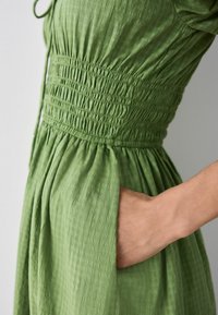 Vihreä mekko, joka on valmistettu kevyestä kankaasta, ja siinä on kuminauharesori vyötäröllä, sivutaskut sekä virtaava siluetti, jossa on tekstuuri.
