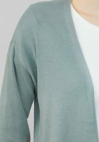 Helle grüne Strickjacke mit geripptem Muster, die einen tiefen V-Ausschnitt und lockere Ärmel hat, über einem weißen, enganliegenden Shirt getragen.