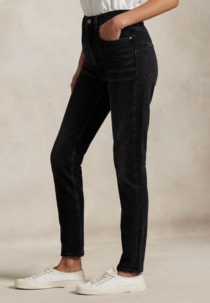 Personne portant un jean noir taille haute et des baskets blanches, debout avec une main dans la poche devant un fond uni.